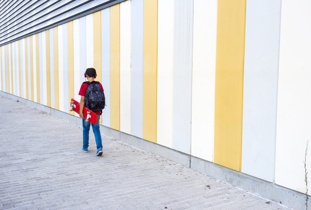 view of a young boy wearing casual clothes walking against a colored wall while holding a skateboard